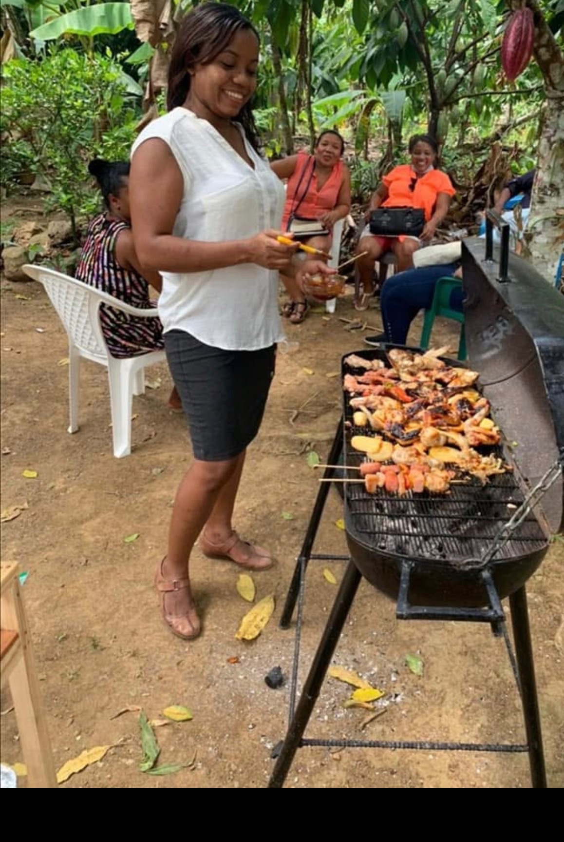 Woman grilling skewers of chicken and vegetables outdoors in the Dominican Republic, friends relaxing in the background under tropical trees