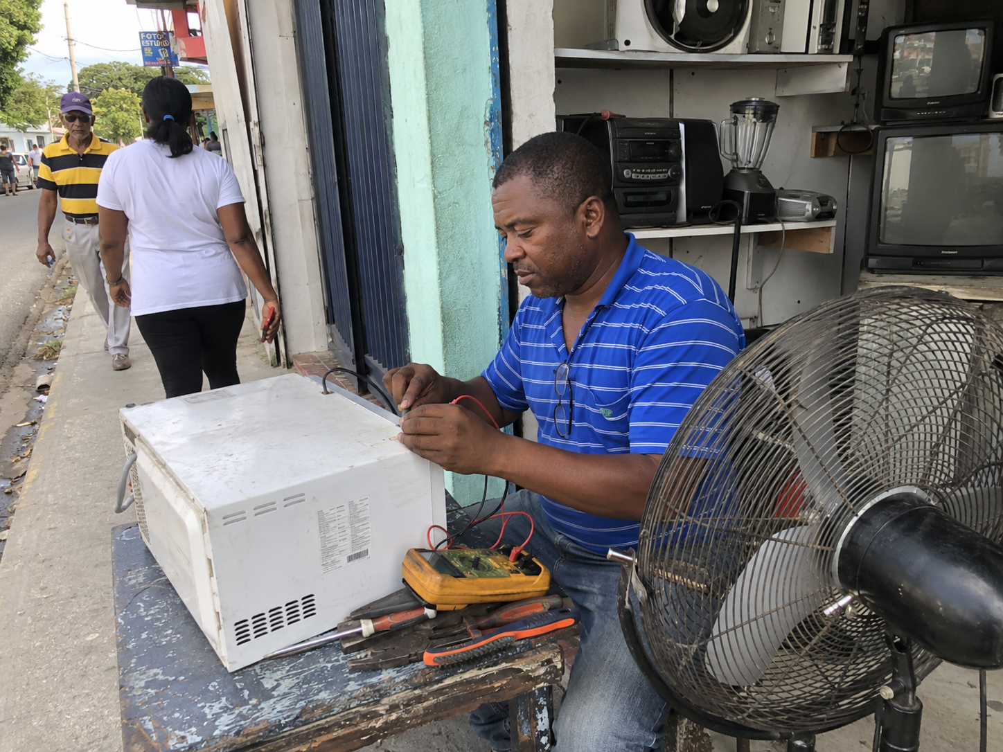 Man repairing a microwave on a sidewalk in the Dominican Republic, TVs and electronics stacked behind him, neighbors walking past