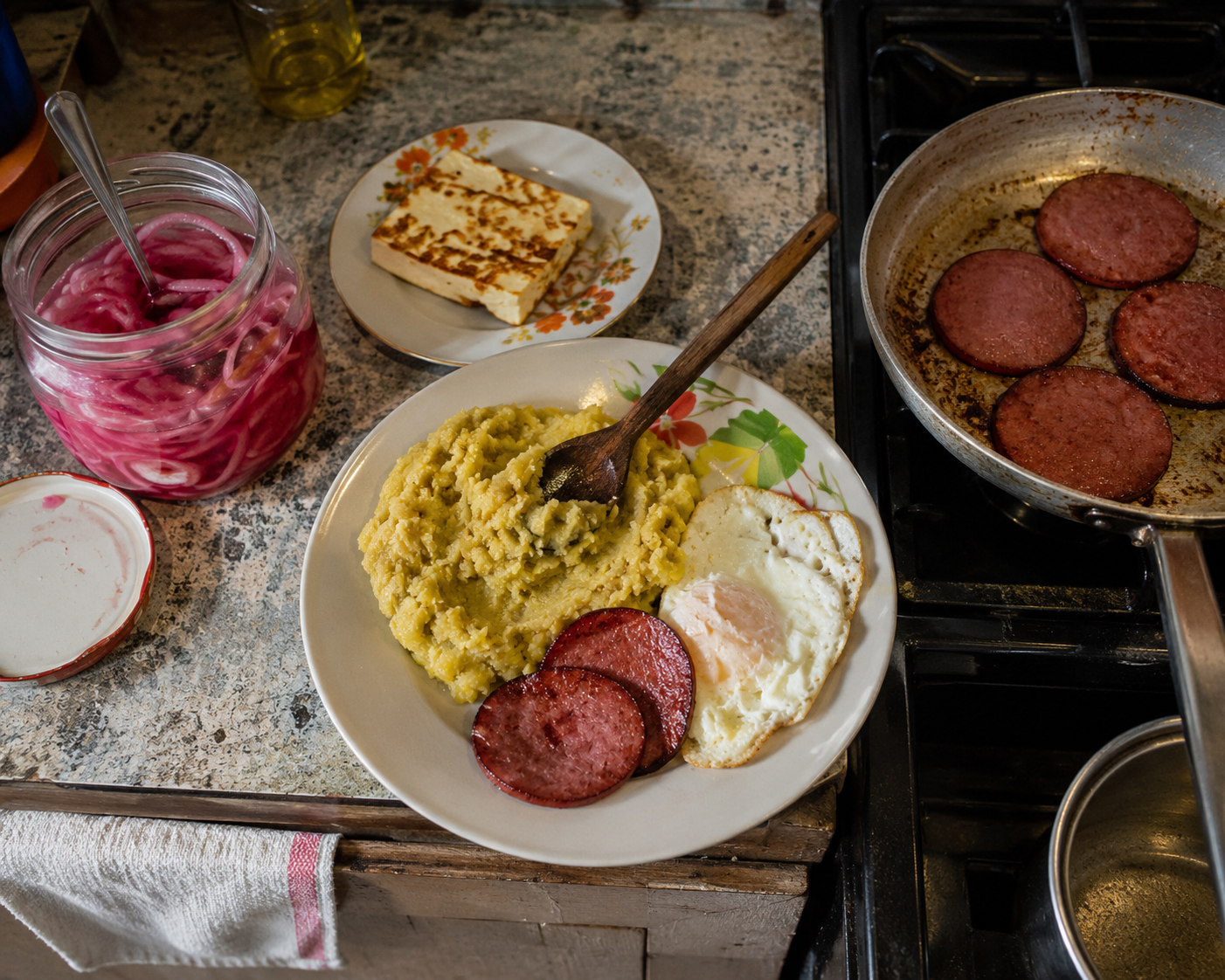 Salami frying in a pan on the stove next to a finished plate of mangu, fried cheese, and eggs on the kitchen counter