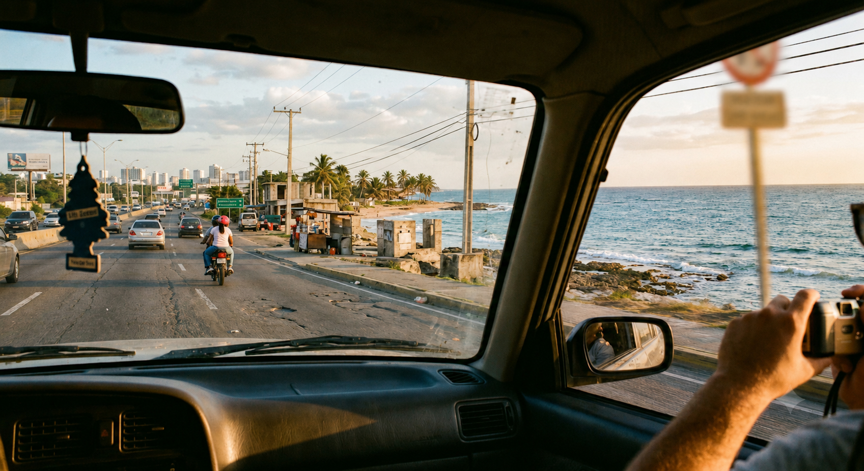 View through a car windshield driving Las Américas Highway toward Boca Chica, motorbike ahead in traffic, roadside vendor stands, and the Caribbean coastline out the passenger window