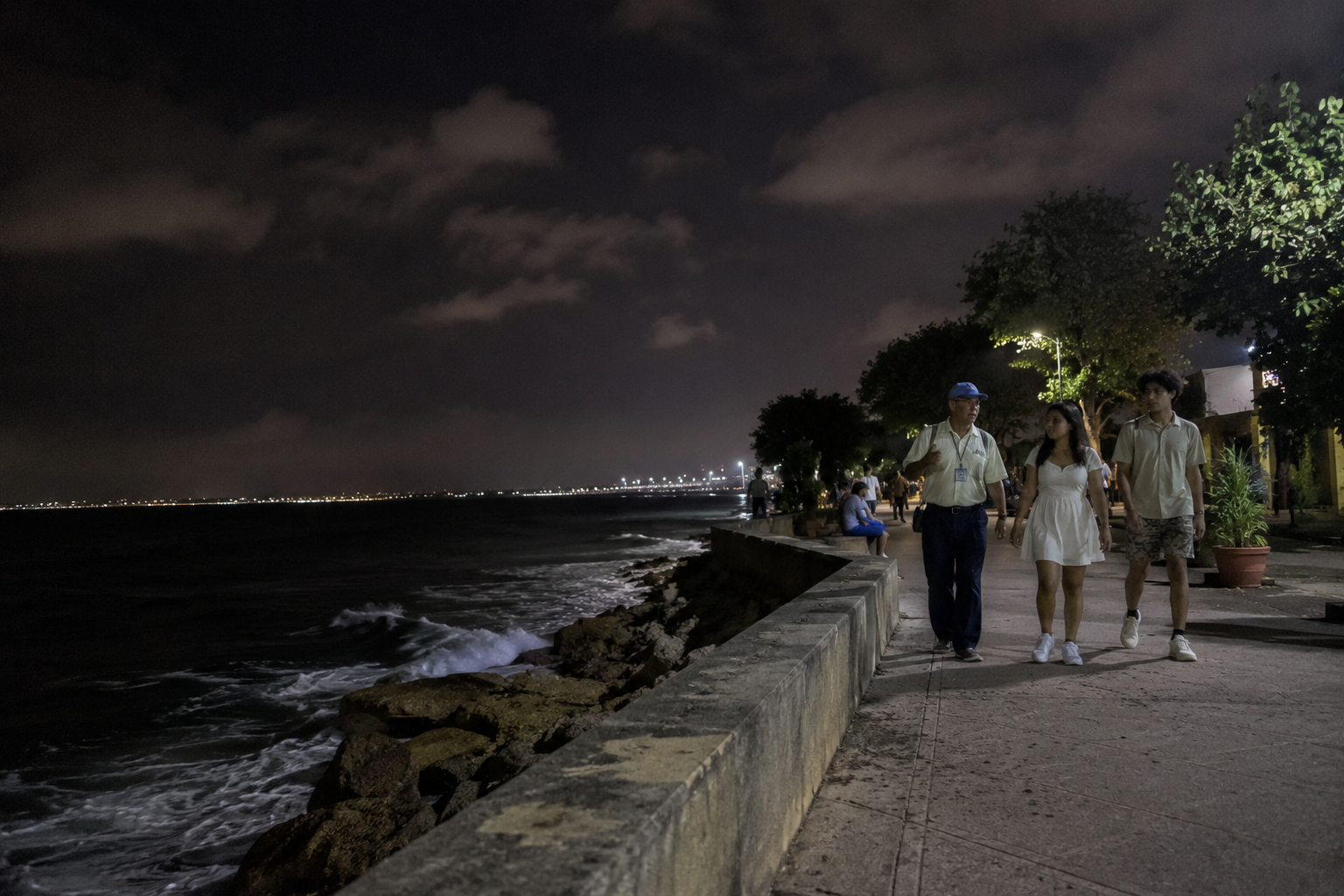 People strolling the Malecón seawall in Santo Domingo at night, waves breaking on the rocks below and city lights stretching down the coast