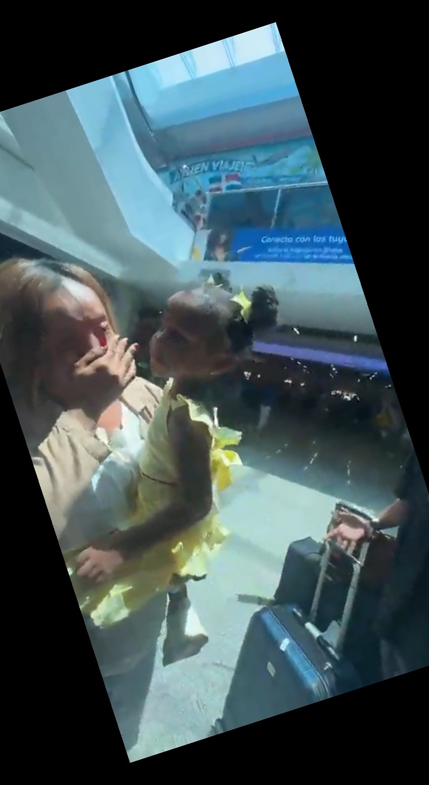 Dorka covering her mouth in joyful shock, holding a child in a yellow dress, DR airport signage visible behind them at SDQ arrivals